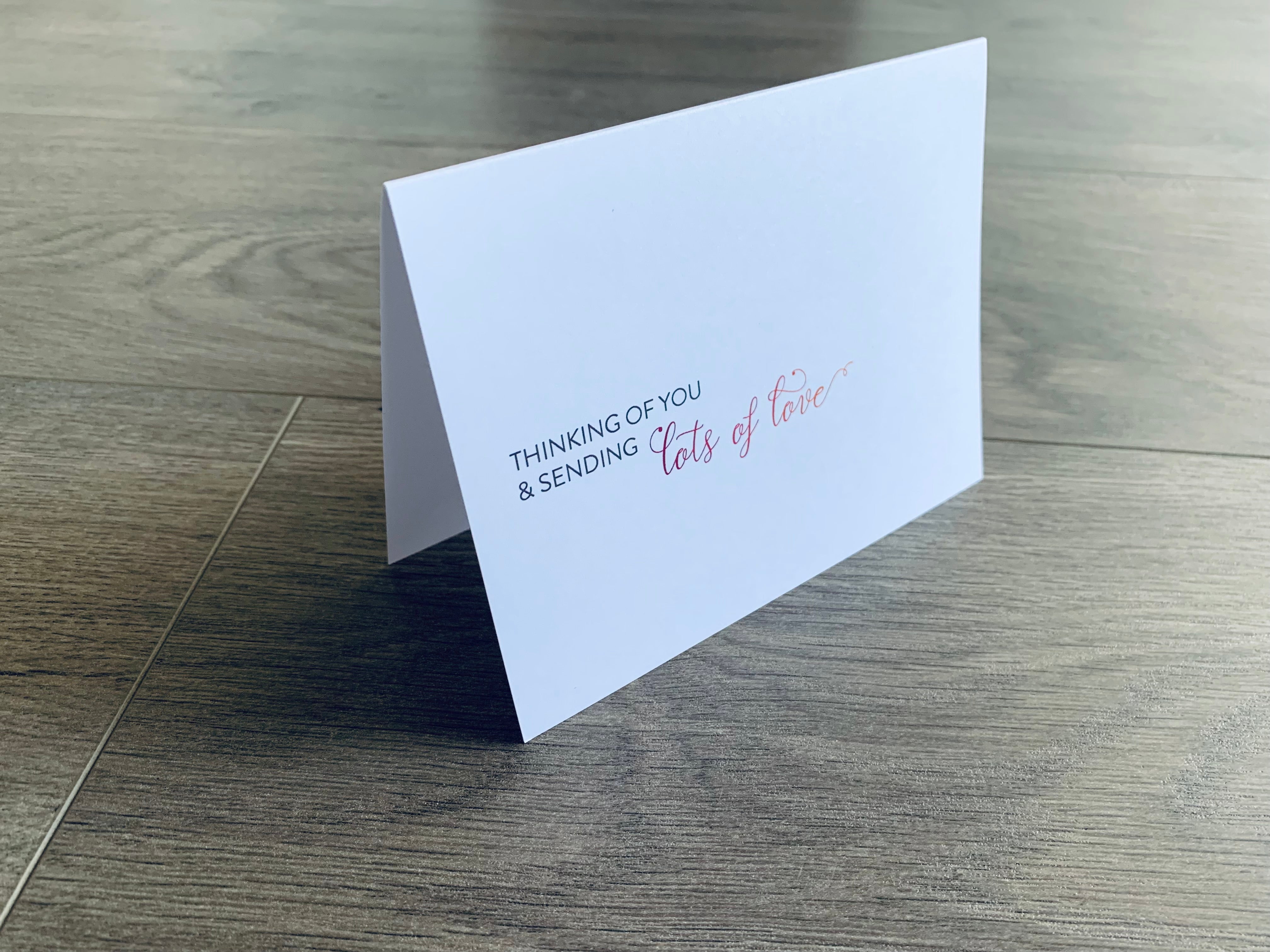 A white notecard is propped up on a gray wooden floor. The card reads, "Thinking of you and sending lots of love." Stationare's Thinking of You collection.