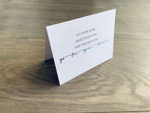 A white folded notecard propped up on a wooden floor. The card reads, "Set your goal. Make your plan. Take the first step. Go for your dream." The final line - go for your dream - is in a wide cursive font that fades in color from purple to aqua. From Stationare's Inspire Collection.