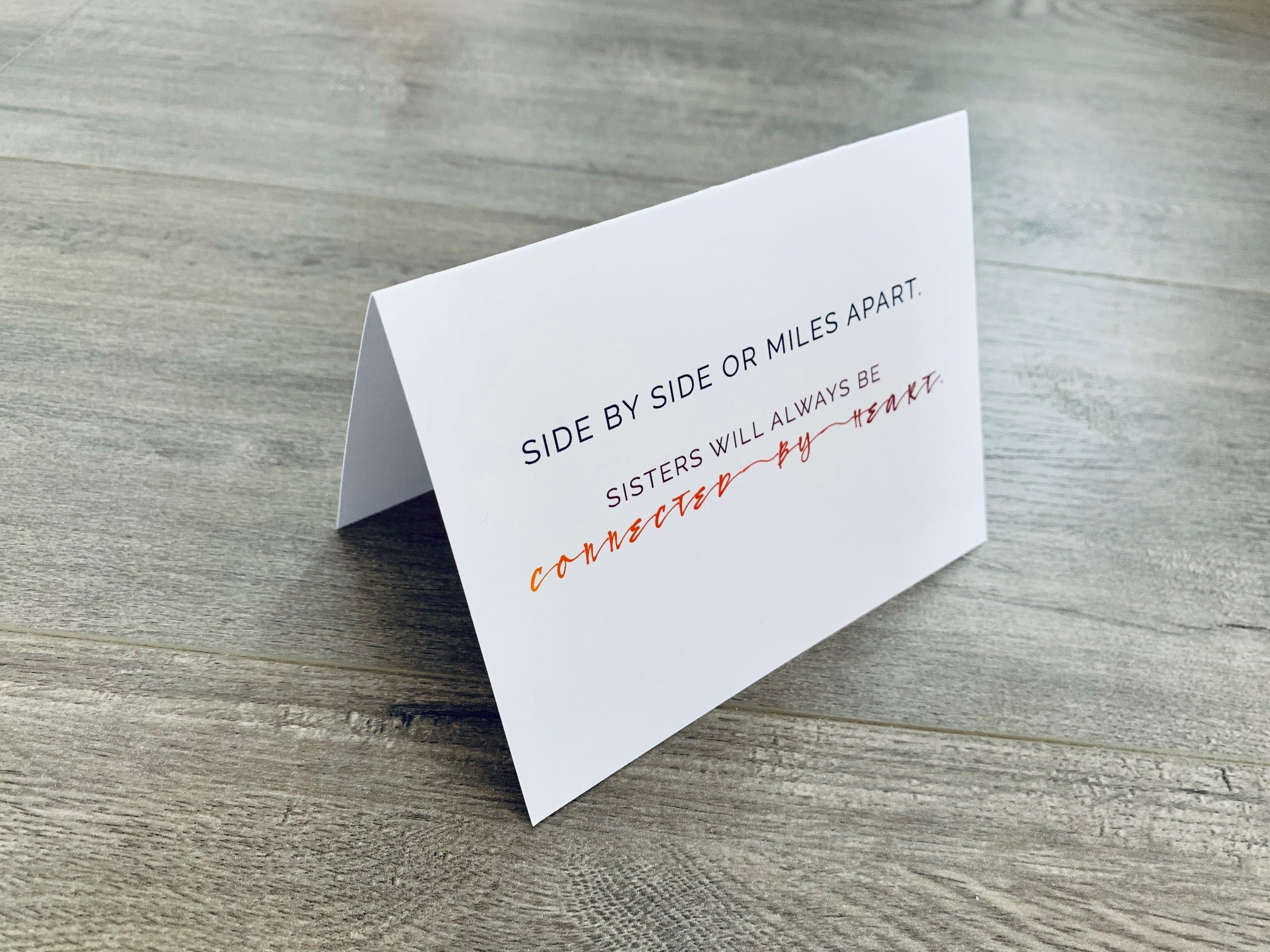 A folded white notecard is propped up on a wooden floor. The card reads, "Side by side or miles apart. Sisters will always be connected by heart." Stationare's Sisters collection.