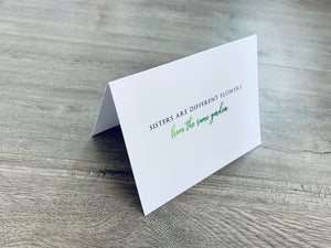 A folded white notecard is propped up on a wooden floor. The card reads, "Sisters are different flowers from the same garden." Stationare's Sisters collection.