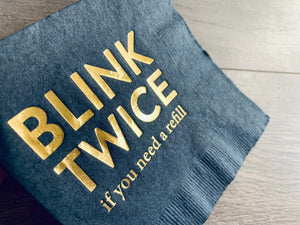 Closeup of a rich black cocktail napkin with gold foil printing that reads, "Blink Twice if you need a refill." By Stationare.