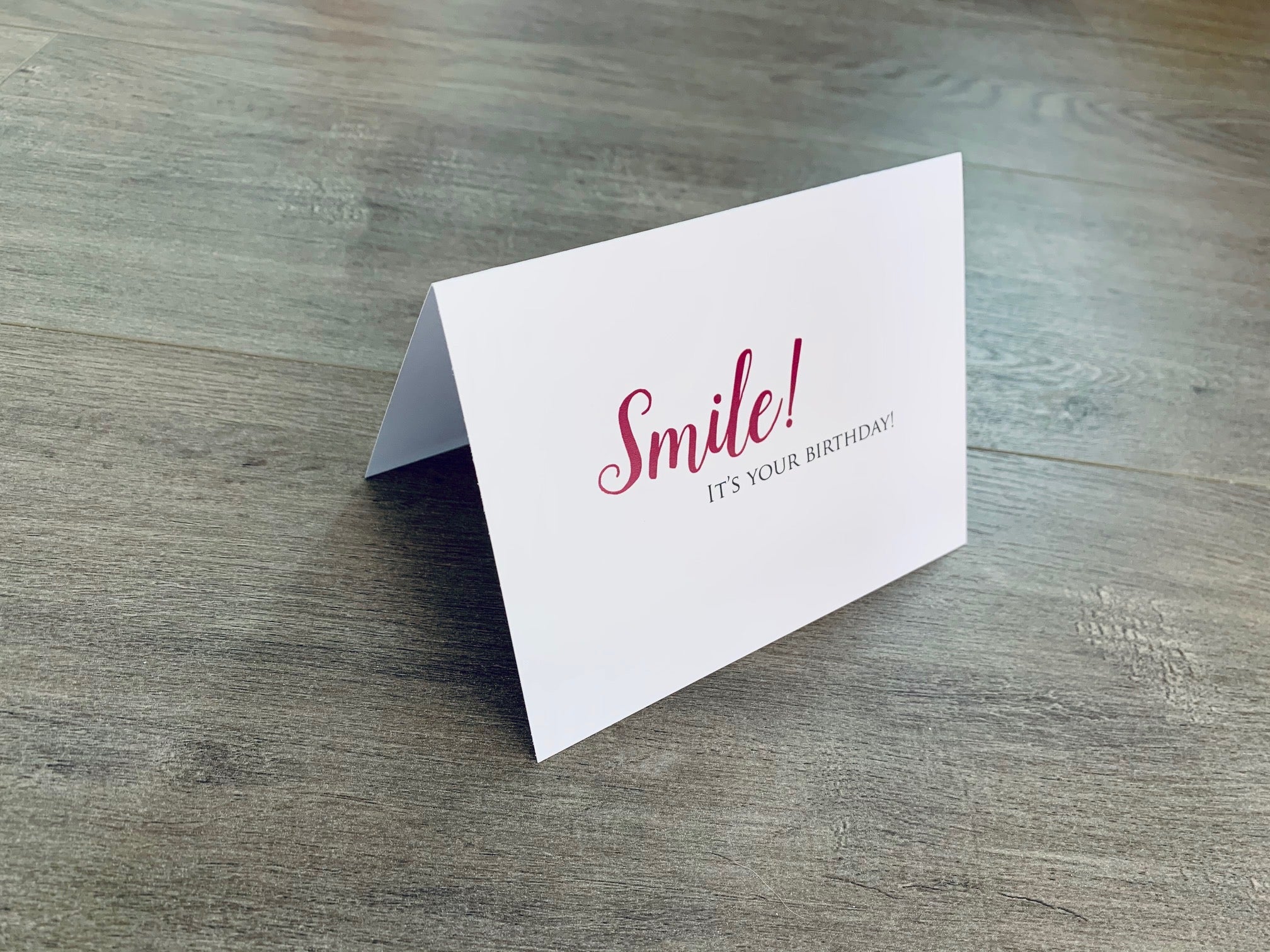 A white card is propped on a gray wood floor. The card reads, "Smile! It's your birthday!" Birthday card by Stationare