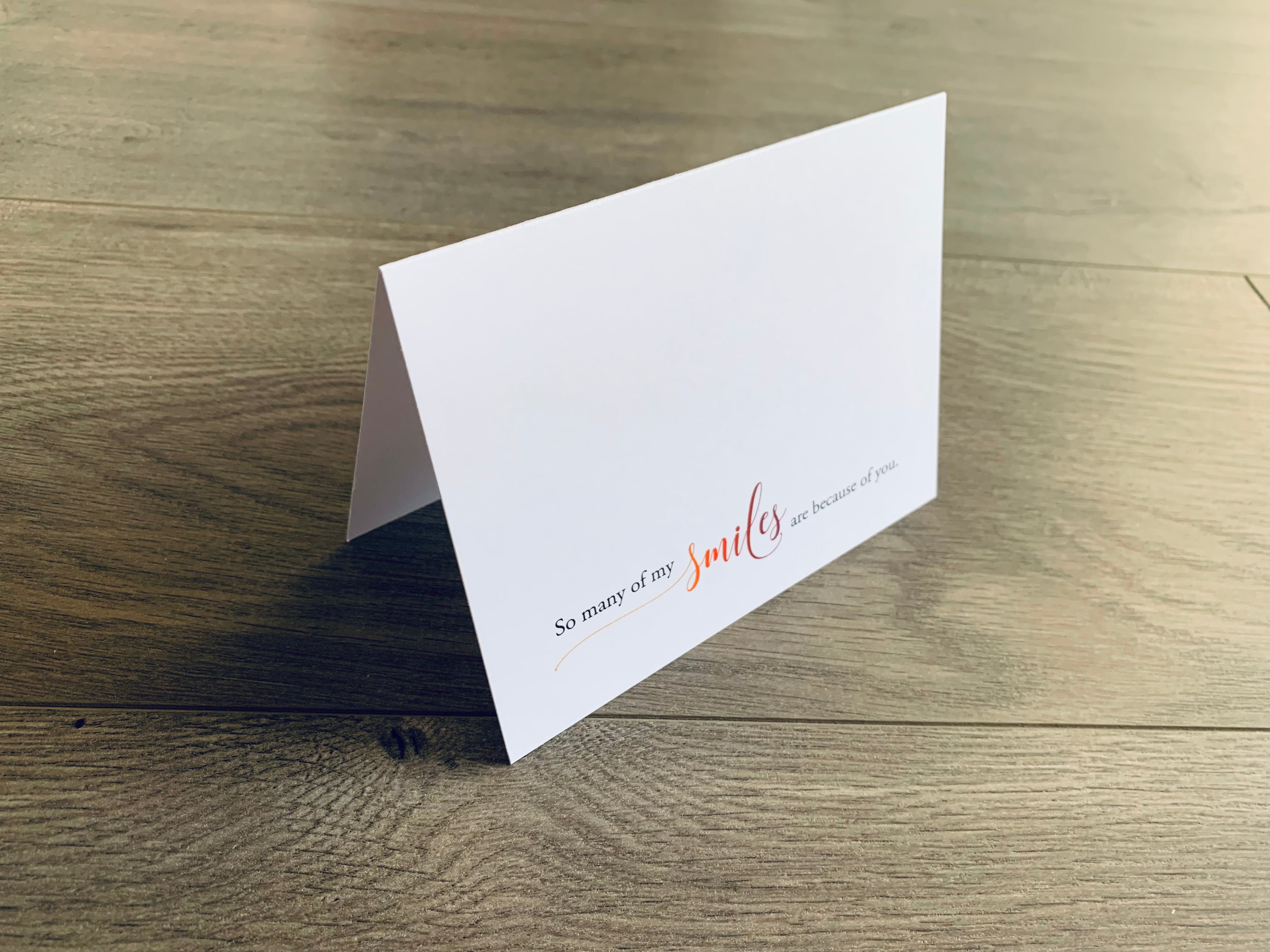 A folded white notecard is propped up on a wooden floor. The card reads, "so many of my smiles are because of you" in a mix of serif and script fonts. Love Collection by Stationare.