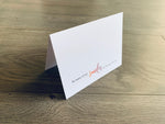 A folded white notecard is propped up on a wooden floor. The card reads, "so many of my smiles are because of you" in a mix of serif and script fonts. Love Collection by Stationare.