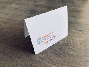 A white, folded notecard is propped up on a gray wooden background. The card says, "It's the strangest thing - sometimes, I open my mouth and I hear my mother." Mother's Day card by Stationare.