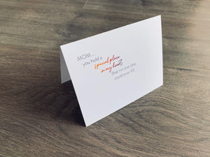 A white, folded Mother's Day card is propped up on a gray wooden floor. The card reads, "Mom... you hold a special place in my heart that no one else could ever fill." Mother's Day collection by Stationare.