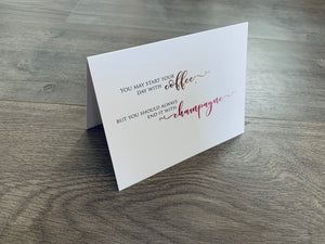 A folded white notecard stands on a gray wooden floor. It reads, "You may start your day with coffee, but you should always end it with champagne." Stationare's Wine and Champagne Lovers Collection.