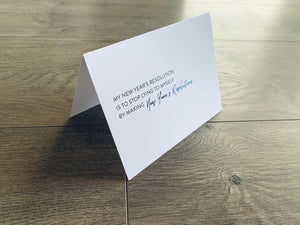 A white folded notecard stands on a gray wood floor. The card reads, "My New Year's resolution is to stop lying to myself by making New Year's Resolutions."