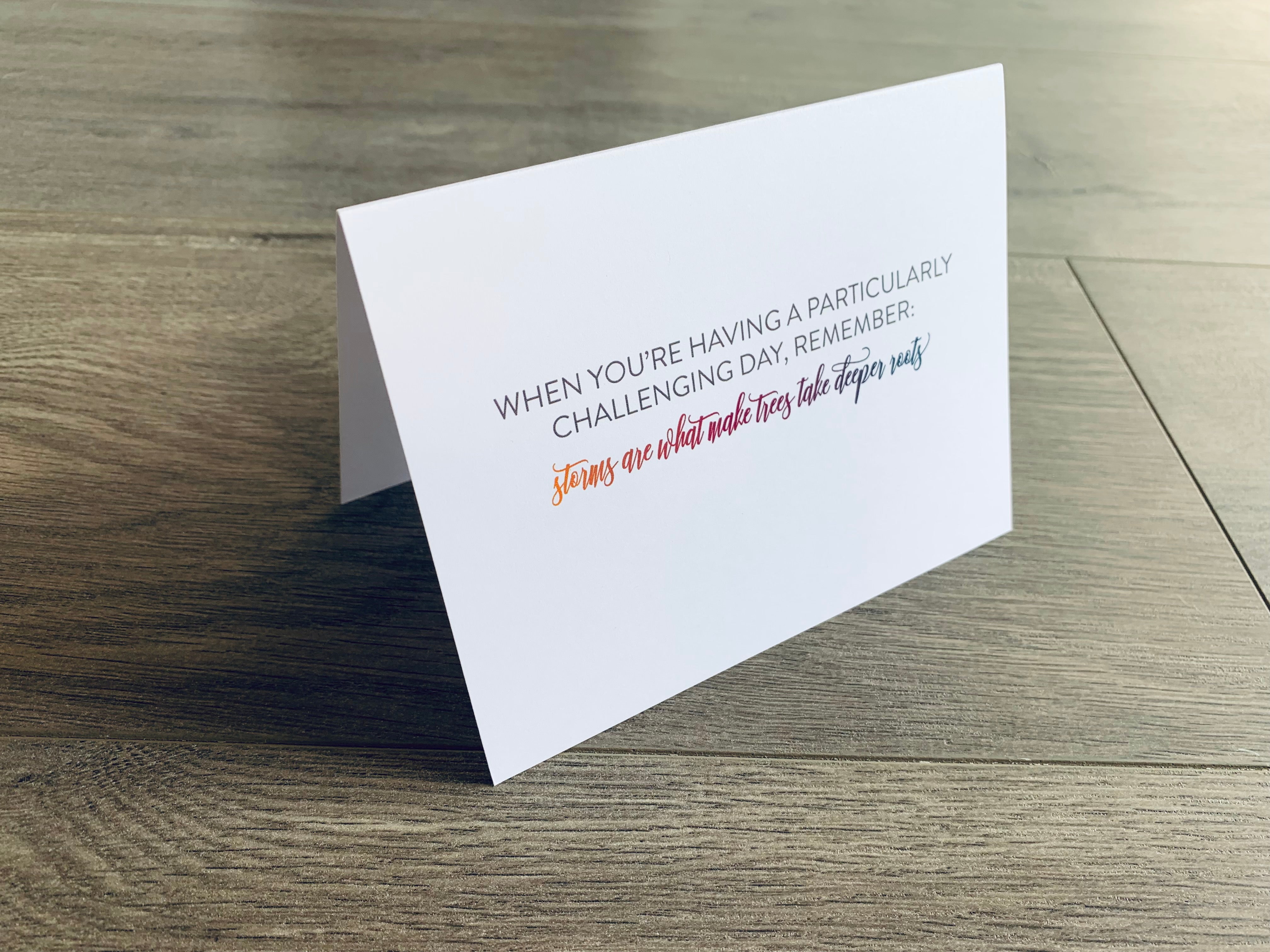 A white, folded notecard sits on a gray wooden floor. The card reads, "When you're having a particularly challenging day, remember: storms are what make trees take deeper roots." Sending Support collection by Stationare.