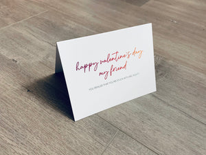 A folded white notecard is folded and propped up on a gray wooden background. The card says, "Happy Valentine's Day my friend... you realize that you're stuck with me, right?" Valentine Smiles collection by Stationare.