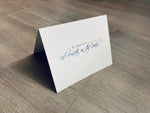 A white card is propped on a gray wood floor. The card reads, "All I need is you and sunsets on the beach." Beach Love Collection by Stationare.