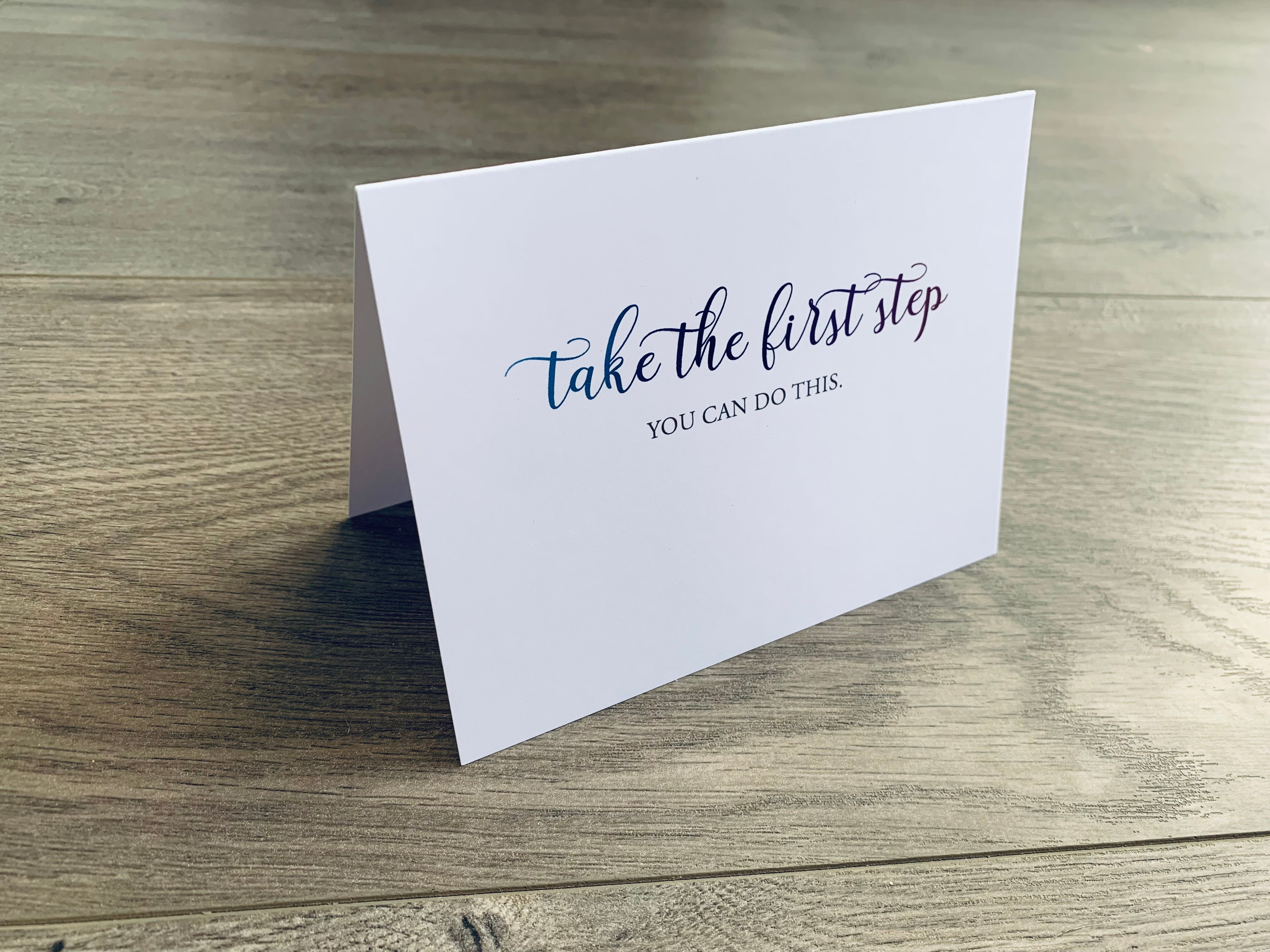 A propped up, white folded notecard sits on a wooden floor. The words "take the first step" are in a script font across the top with the wording - you can do this - below it. From Stationare's Inspire Collection.