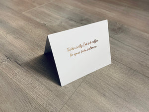 A white, folded notecard sits on a wooden floor. The card says, "Technically, I drink coffee for your sake, not mine." Coffee and Conquer collection by Stationare.