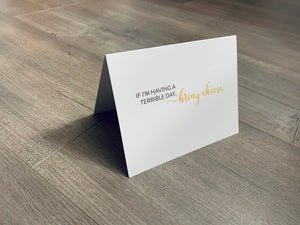 A white notecard is on a gray wooden floor. The card says, "If I'm having a terrible day, bring cheese." Cheese Lovers collection by Stationare.