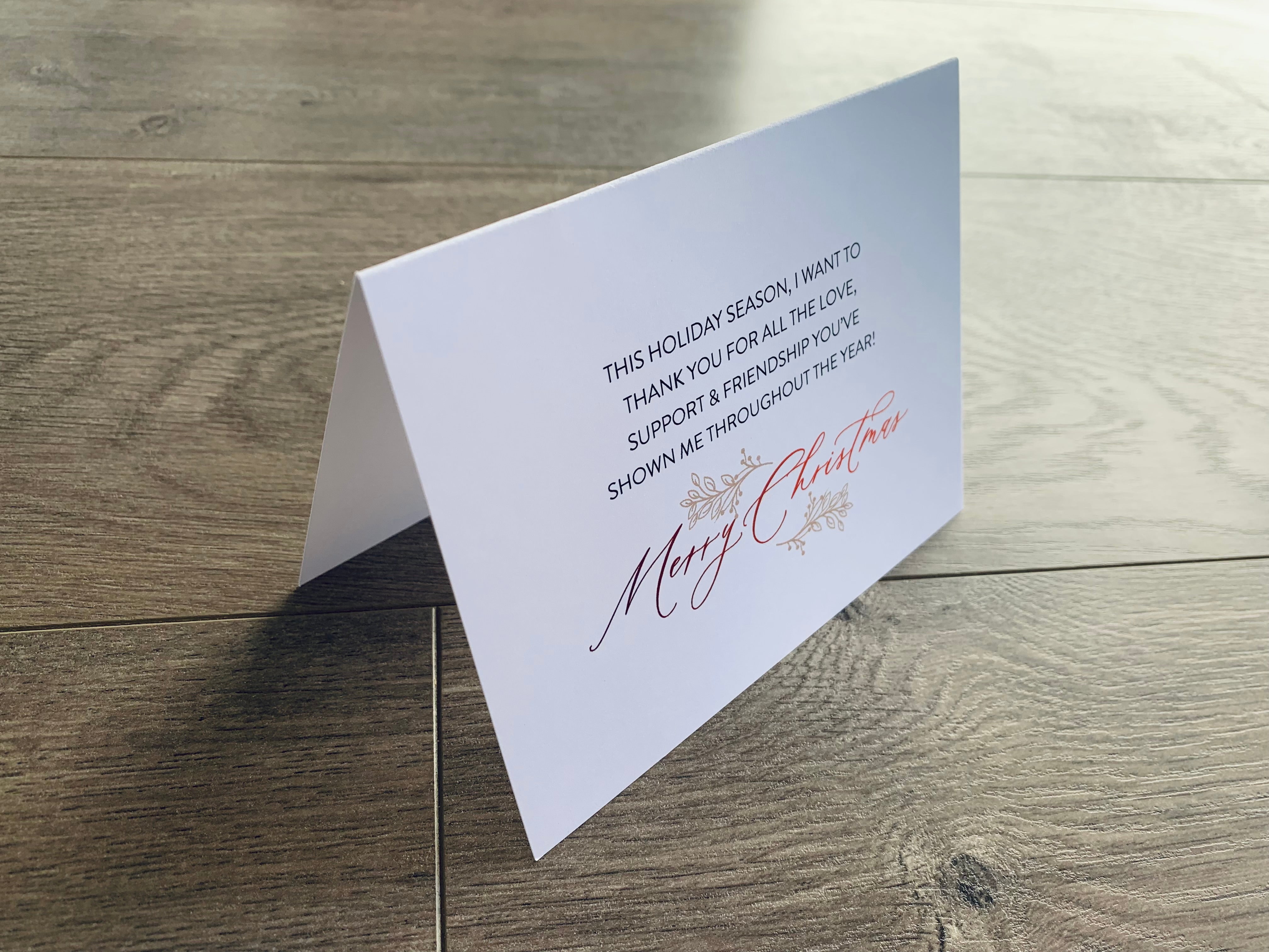 A white folded notecard sits on a wooden floor. On the front of the card, it reads, "This holiday season, I want to thank you for all the love, support & friendship you've shown me throughout the year! Merry Christmas!" Merry Thanks Collection by Stationare.