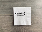 White cocktail napkin lies on a gray wooden floor. In brown matte foil it reads, "Thankful for cheese & wine" with five small leaves.
