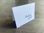 A white notecard is propped up on a gray wooden floor. The card reads, "Thinking of You" in a teal script font. Thinking of You Collection by Stationare.
