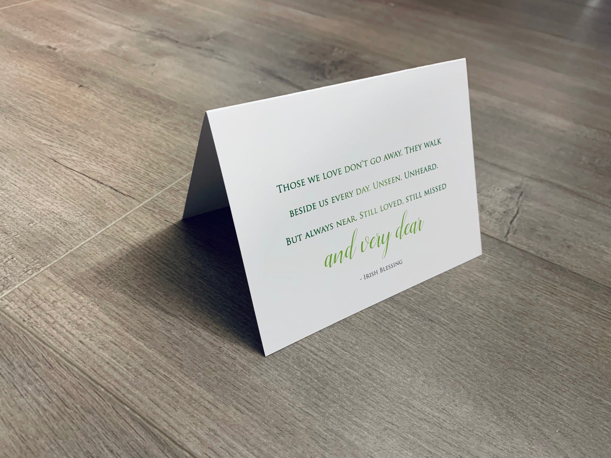 A white folded notecard is propped up on a gray wooden floor. In green printing, it reads, "Those we love don't go away. They walk beside us every day. Unseen. Unheard. But always near. Still loved. Still Missed and very dear. - Irish Blessing." From Stationare's In Sympathy collection.