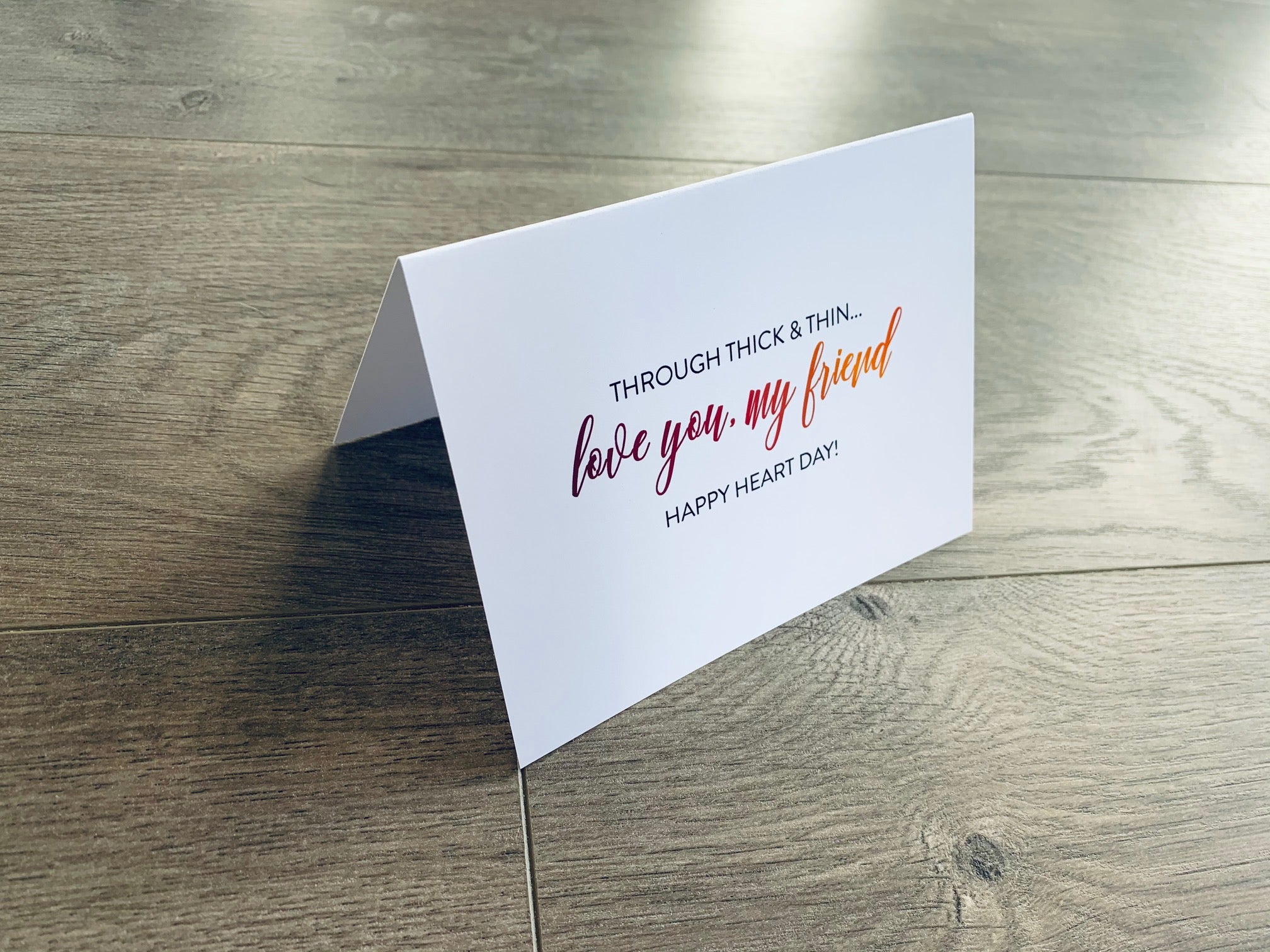 A folded white notecard is folded and propped up on a gray wooden background. The card says, "Through thick and thin... love you, my friend. Happy Heart Day!" VDay BFFs collection by Stationare.