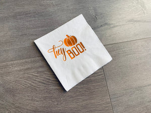 White tilted cocktail napkin on a gray wooden background. The napkin reads, "hey Boo!" with a small pumpkin in shiny orange foil.