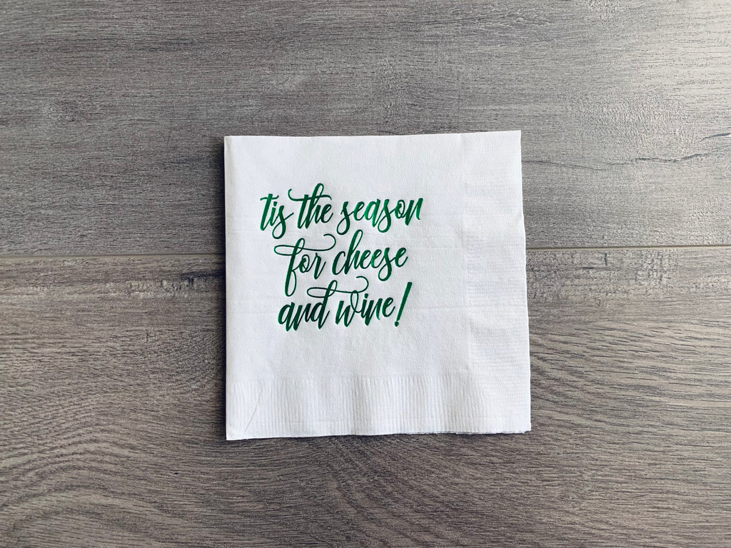 White cocktail napkin sits on a gray wooden background. In metallic green foil, it reads: "tis the season for cheese and wine!" By Stationare