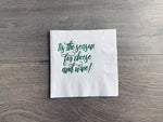 White cocktail napkin sits on a gray wooden background. In metallic green foil, it reads: "tis the season for cheese and wine!" By Stationare