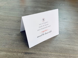 A white, folded notecard sits on a wooden floor. The card says, "lots of laughter, family and friends, and a homemade meal... together... around the dinner table." The Foodie collection by Stationare.