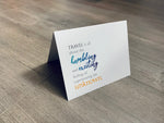 A folded white notecard is propped up on a wooden floor. The card reads, "Travel is all about the humbling and exciting feeling of experiencing the unknown." Stationare's Travel Lovers collection.