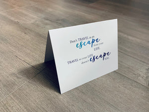 A folded white notecard is propped up on a wooden floor. The card reads, "Don't travel as an escape from the life. Travel so your life doesn't escape you." Stationare's Travel Lovers collection.