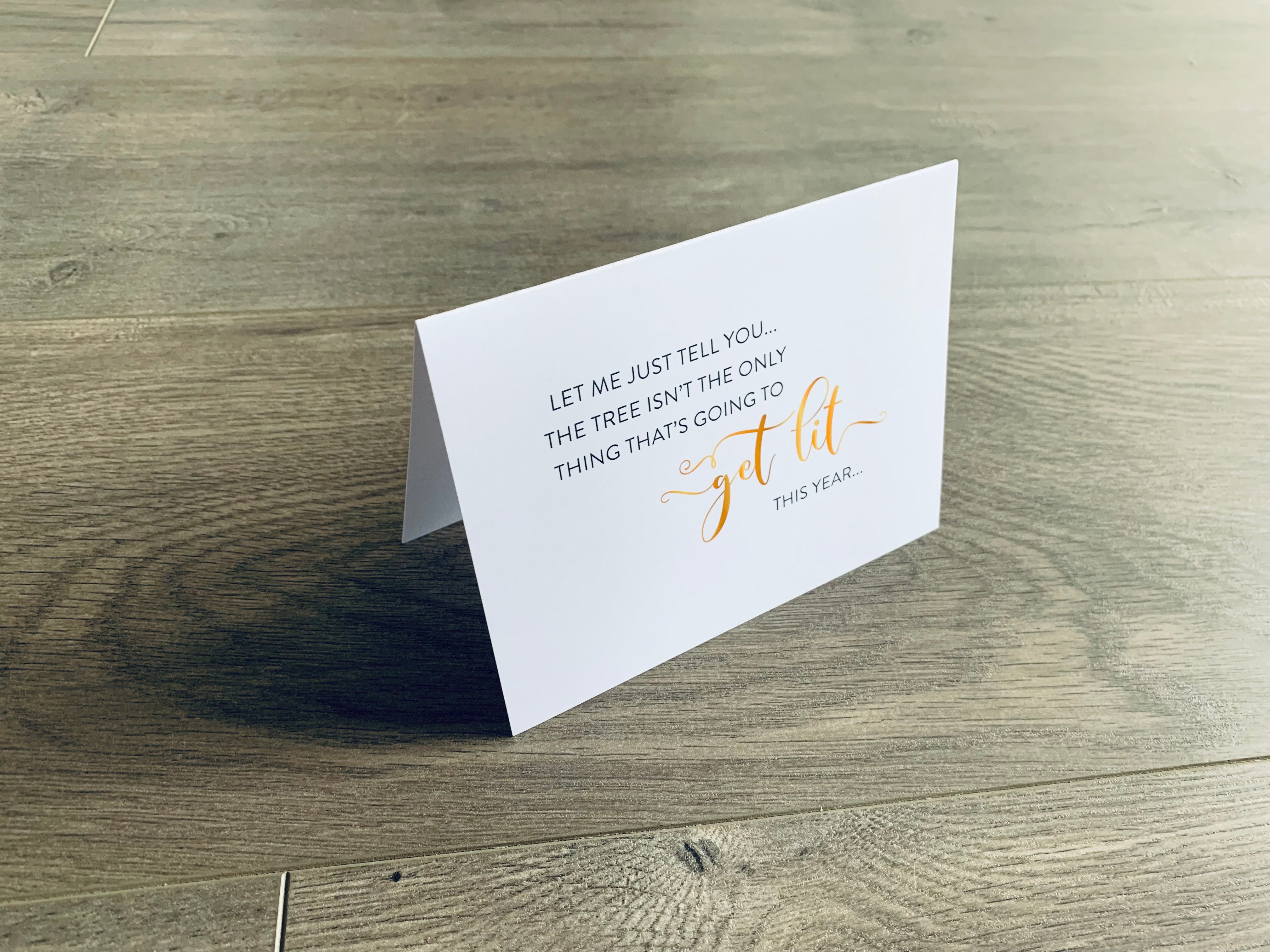 A white folded notecard is propped up on a wooden floor. On the front of the card, it says "Let me just tell you... the tree isn't the only thing that's going to get lit this year." Christmas Chuckles collection by Stationare.