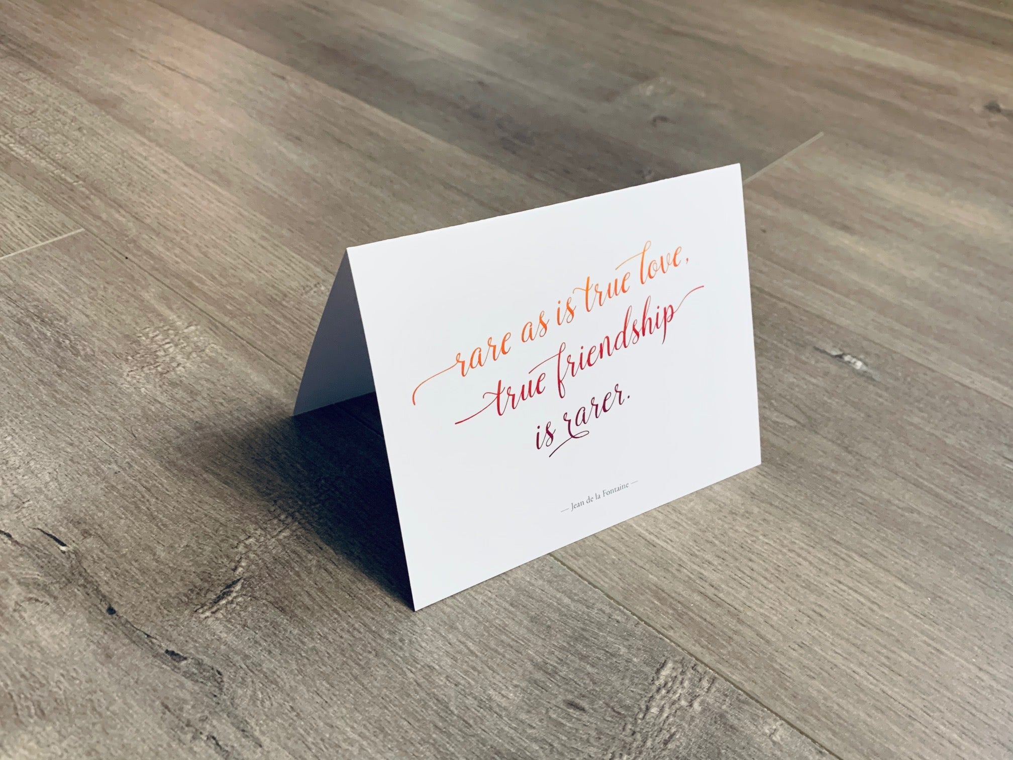 A white, folded notecard sits on a wooden floor. The card has a quote by Jean de la Fontaine and says, "rare as is true love. true friendship is rarer." The Friendship Valentines collection by Stationare.