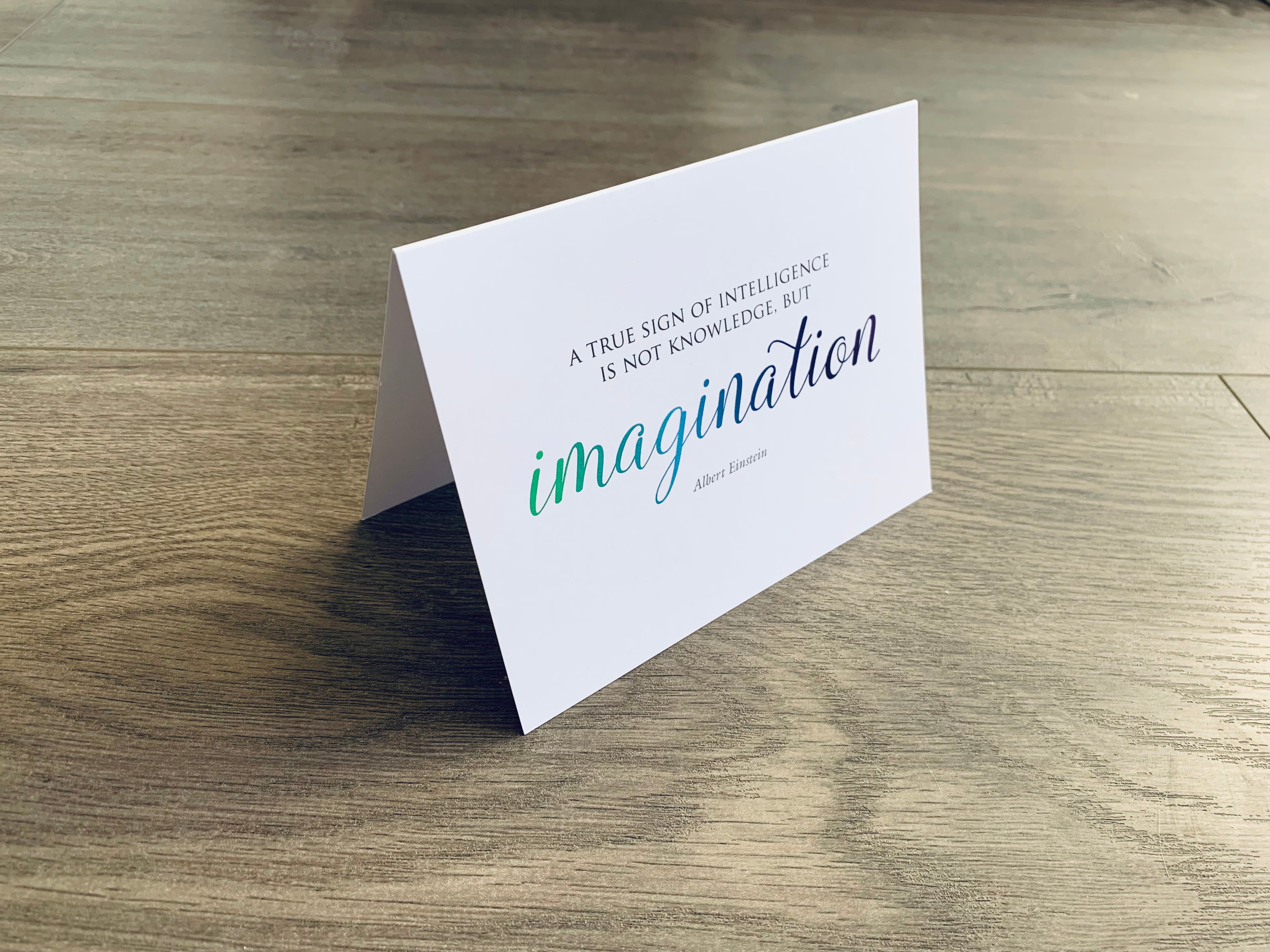 A white, folded notecard sits on a wooden floor. The card says, "A true sign of intelligence is not knowledge, but imagination." Creativity collection by Stationare.
