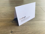 A white notecard is folded and standing on a wooden floor. The card reads "True strength comes in the ability to break a chocolate bar into four equal pieces with your bare hands...and then only eat one of those pieces." Card by Stationare.
