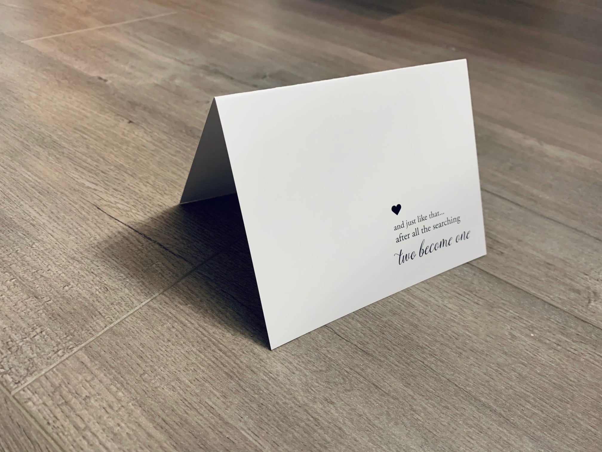 A white notecard with black ink is folded and propped up on a wooden floor. The card says, "and just like that... after all the searching two become one." Bridal and Wedding collection by Stationare.