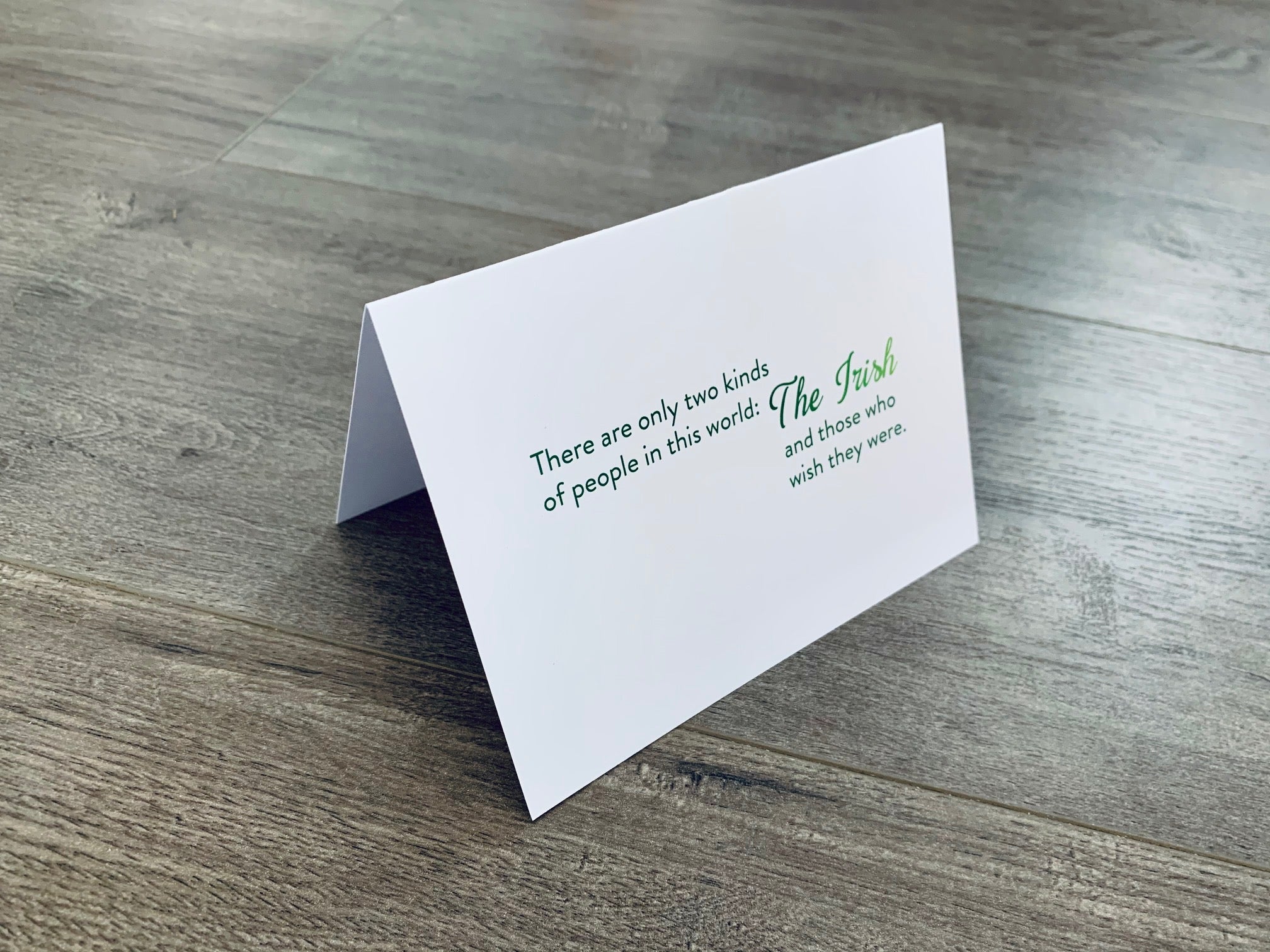 A white folded notecard is propped up on a wooden floor. The card has the Irish proverb, "There are only two kinds of people in this world - The Irish and those who wish they were." From the Irish Laughs Collection by Stationare.