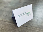 A white folded notecard is propped up on a wooden floor. The card has the Irish proverb, "There are only two kinds of people in this world - The Irish and those who wish they were." From the Irish Laughs Collection by Stationare.