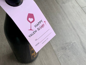 light pink wine bottle tag on a black wine bottle. The tag is light pink with a dark pink wine glass with a light pink heart inside it. The text reads - Happy Valen-wine? with a to and from section to address the recipient.