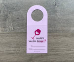 Straight image of the light pink Valen-wine bottle tag. It reads - Happy Valen-wine? with a dark pink wine glass with a light pink heart inside. The tag is lying flat on a gray wood background.