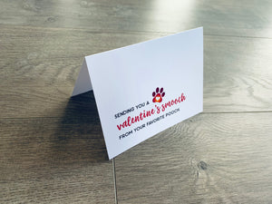A folded white notecard is folded and propped up on a gray wooden background. The card says, "Sending you a Valentine's smooch from your favorite pooch." Valentines from the Pup collection by Stationare.