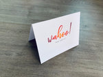 A white, folded notecard sits on a wooden floor. The card says, "Wahoo! You did it!" Congrats collection by Stationare.