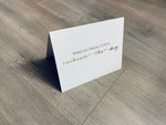 A white, folded notecard sits on a wooden floor. The card says, "Wake up. Drink coffee. Embrace the day." Coffee Lovers collection by Stationare.