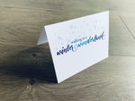 A white, folded notecard sits on a wooden floor. The card says, "walking in a winter wonderland" with small blue snowflakes around the top. Christmas Magic collection by Stationare.