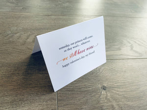 A folded white notecard is folded and propped up on a gray wooden background. The card says, "Someday, our prices will come, or they won't... whatever. We still have wine. Happy Valentine's Day, my friend!" VDay BFFs collection by Stationare.