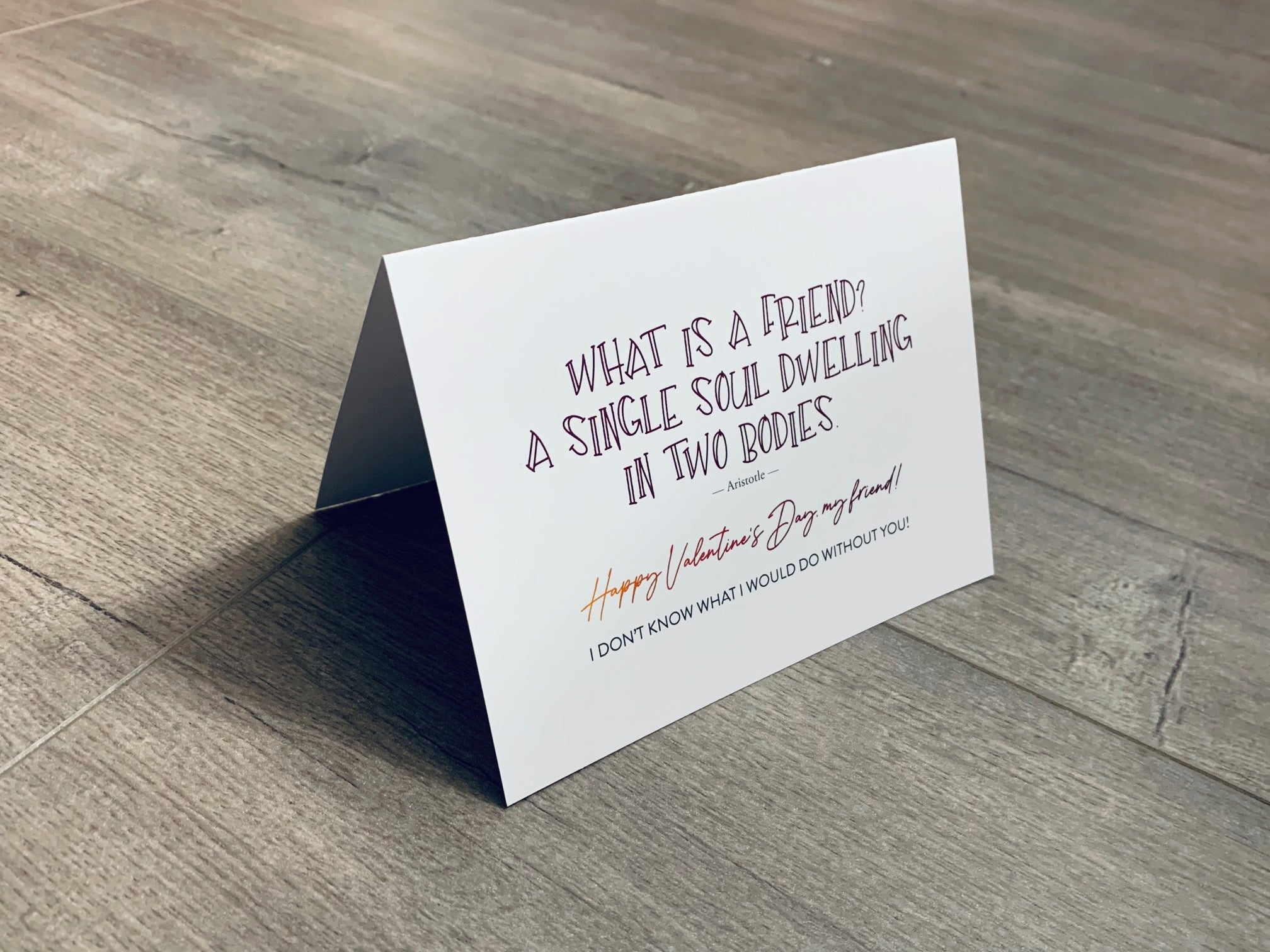 A white, folded notecard sits on a wooden floor. The card has a quote by Aristotle and says, "What is a friend? A single soul dwelling in two bodies." The Friendship Valentines collection by Stationare.