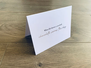 A white card from Stationare's Chocolate Lovers collection is propped up on a wooden floor. The card reads "When life throws a curveball, chocolate saves the day."