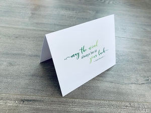 A white folded notecard sits on a gray wood floor. It reads, "may be the wind always be at your back." Irish Inspirations collection by Stationare.