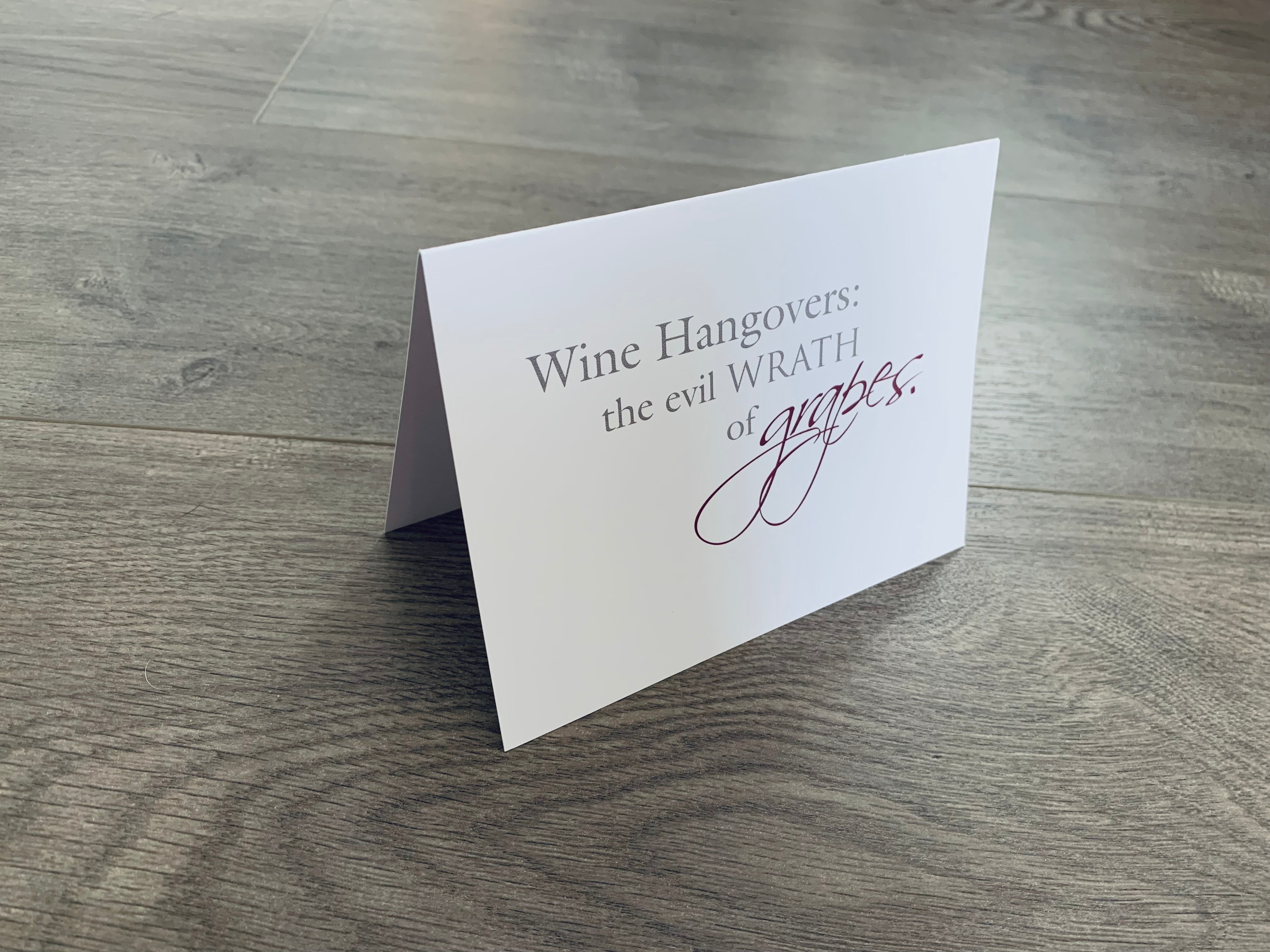 A white notecard from Stationare's Wine and Champagne Lovers' collection is propped up on a gray wooden floor. It reads, "Wine hangovers: the evil wrath of grapes." 