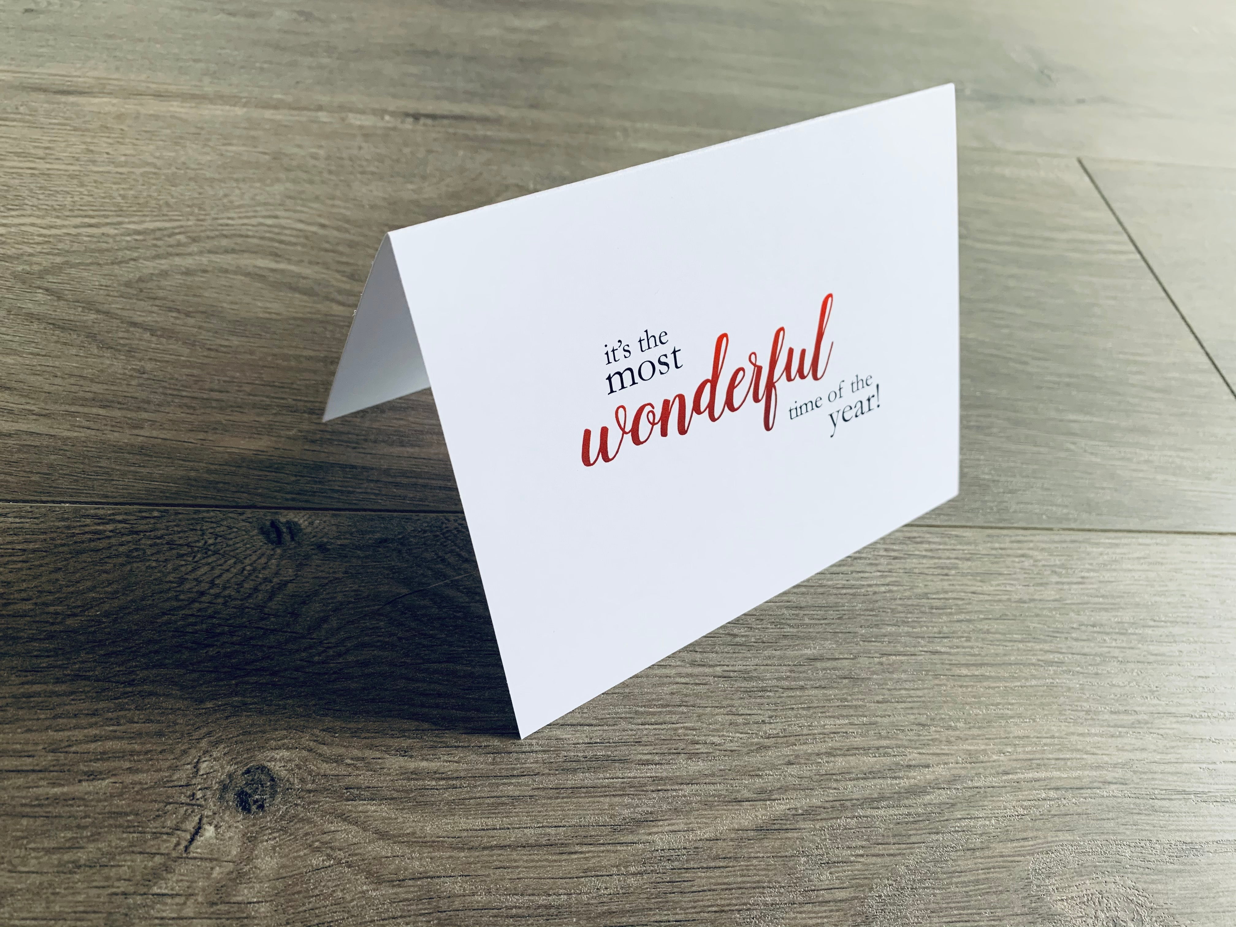A white, folded notecard sits on a wooden floor. The card says, "it's the most wonderful time of the year!" Christmas Magic collection by Stationare.