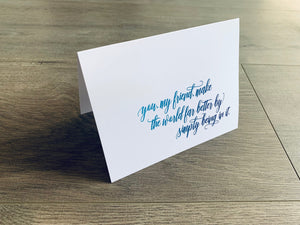 A white, folded notecard sits on a wooden floor. The card says, "you, my friend, make the world far better by simply being in it." The Friendship collection by Stationare.
