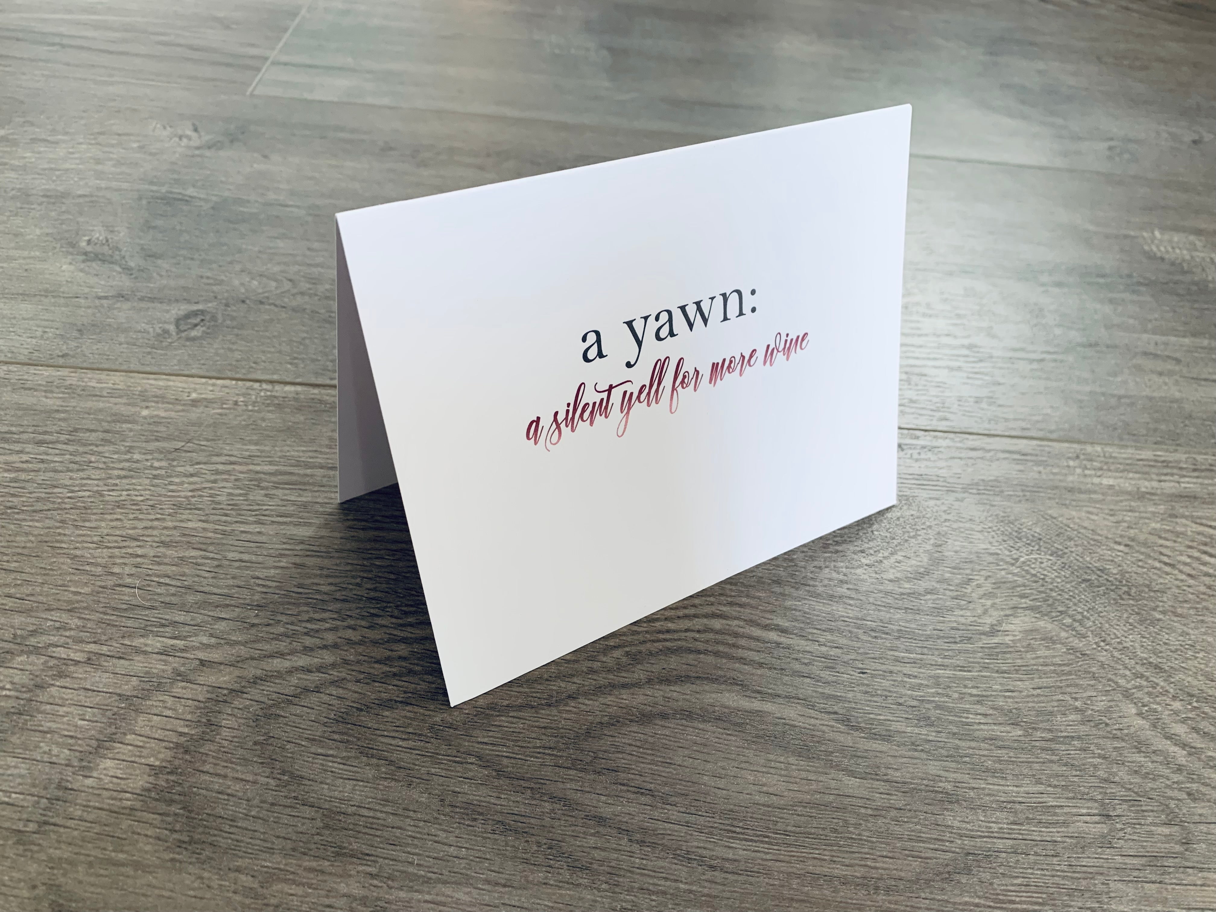 A folded white notecard is propped up on a gray wood floor. The card says, "a yawn: a silent yell for more wine." From Stationare's Wine & Champagne Lovers' Collection.